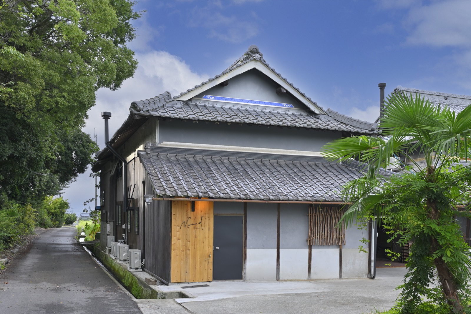 細い道の右手に建つNomad Mannouの外観。瓦屋根の古民家で、正面に杉板の看板と土間がある
