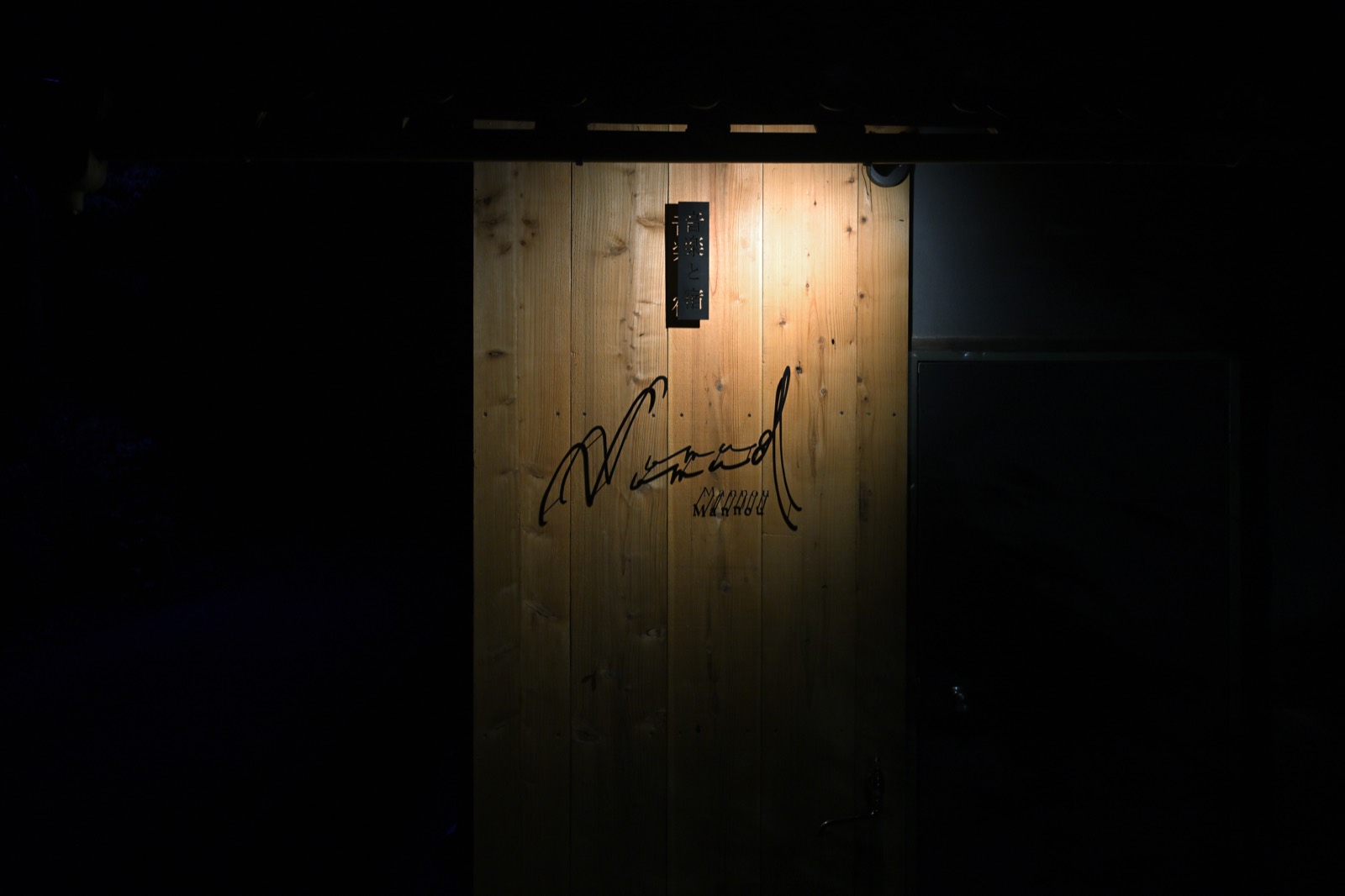 夜のNomad Mannouの看板。杉板がスポットライトで照らされ、サインが浮かび上がっている
